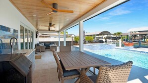 An outdoor patio with a pool, wooden table, wicker chairs, ceiling fans, and a view of the pool area.