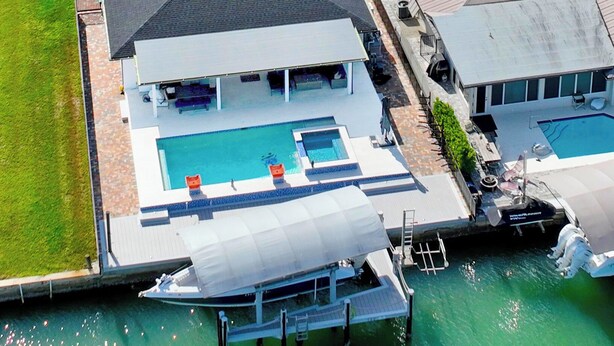A large house with a swimming pool, a boat docked at the pier, and a covered outdoor area.