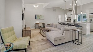 Een moderne woonkamer met een hoekbank, een glazen salontafel en een geel fauteuil. Achtergrond toont een keuken met witte kasten en roestvrijstalen apparatuur.