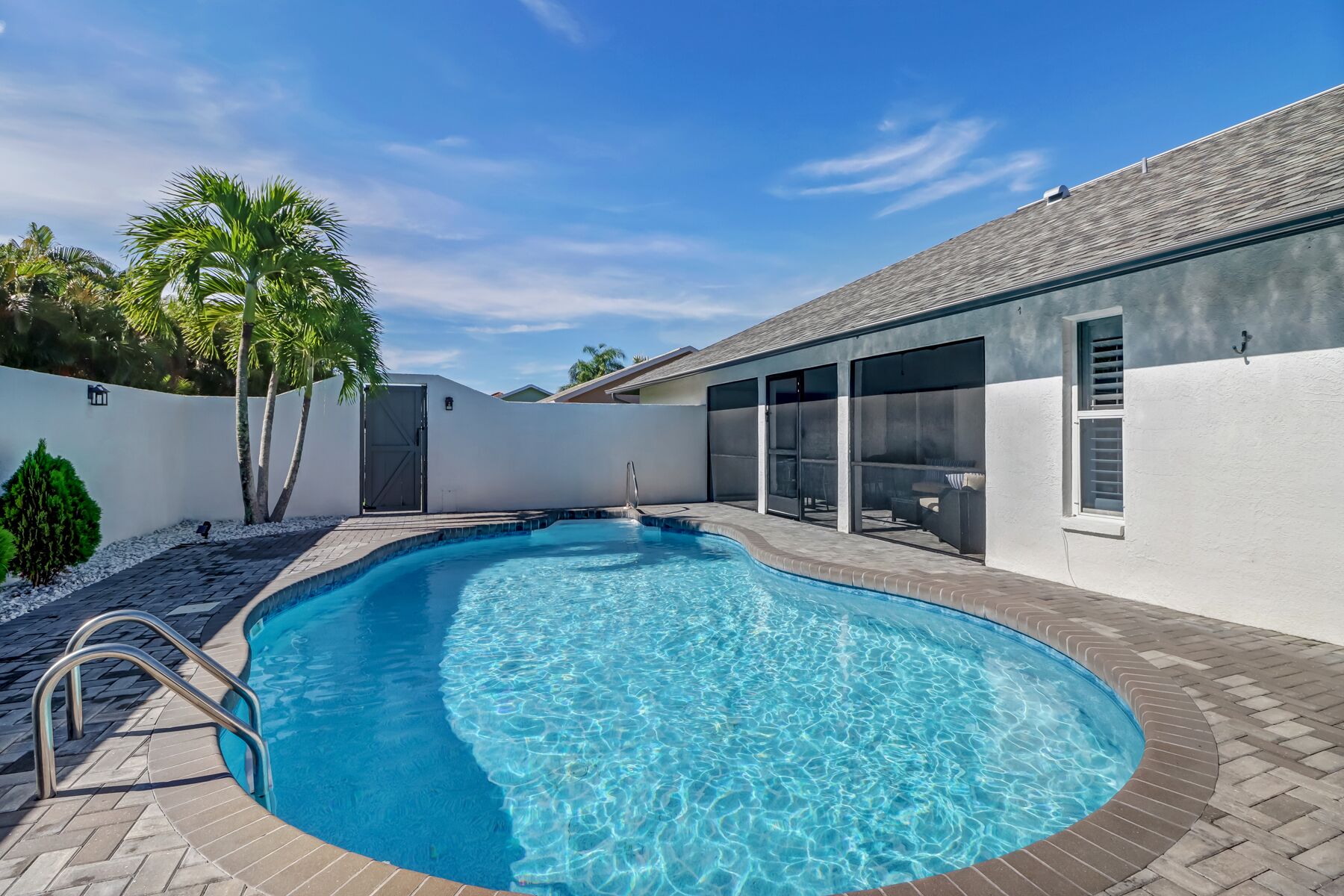 A swimming pool with a brick deck, a covered patio area with sliding glass doors, and a white wall with a door.