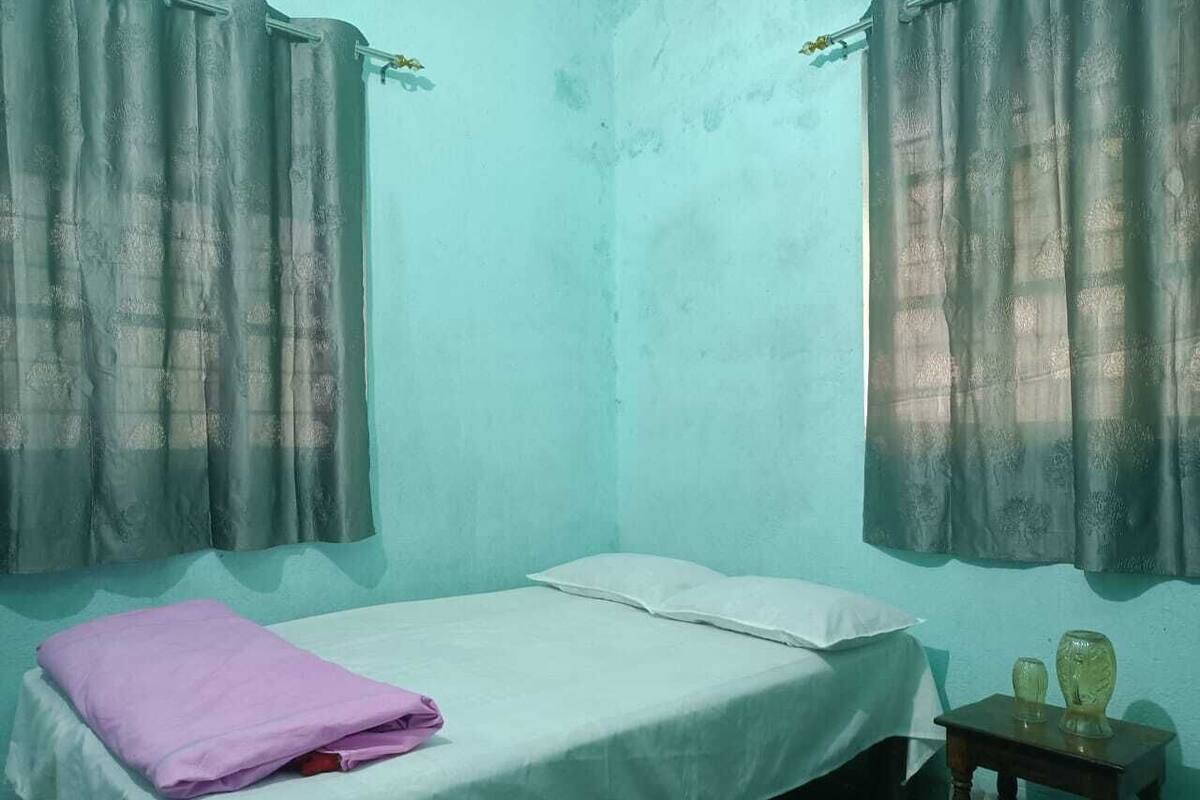A small, single-bed room with a plain bed, a small bedside table, and a window with a curtain.