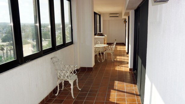 Un pasillo con suelo de baldosas, sillas blancas y una mesa, que conduce a un balcón con vistas a la ciudad.