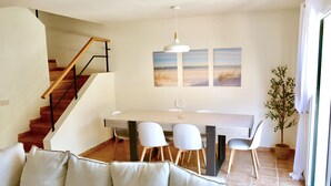 Ein modernes Wohnzimmer mit Sofa, Esstisch mit Stühlen, einer Treppe und Blick auf den Strand durch das Fenster.