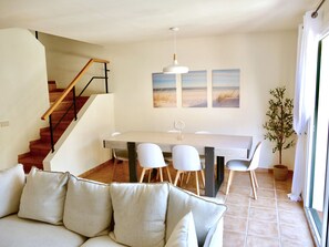 Ein modernes Wohnzimmer mit Sofa, Esstisch mit Stühlen, einer Treppe und Blick auf den Strand durch das Fenster.