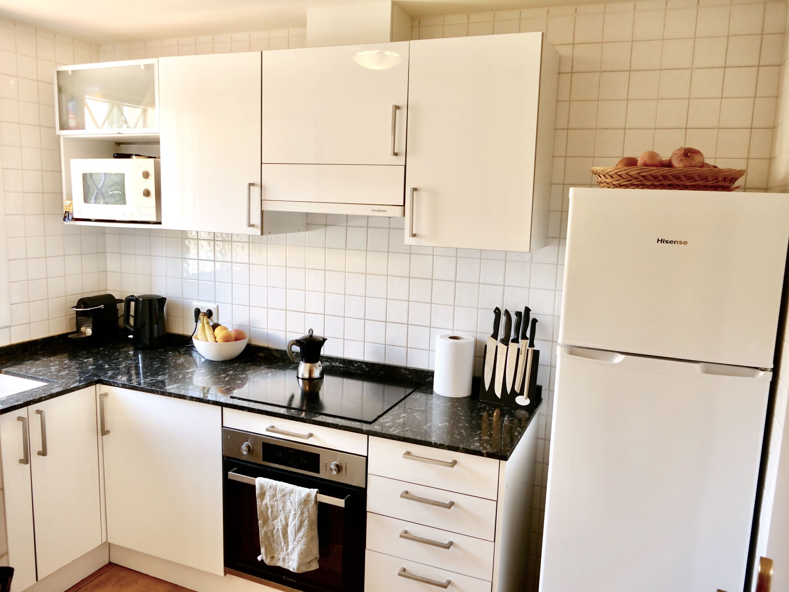A modern kitchen with white cabinets, a black countertop, a Hisense refrigerator, a coffee maker, a kettle, a bowl of fruit, and a knife block.