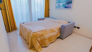 Quarto de hotel com cama, mesa de cabeceira, uma cadeira, um quadro na parede e cortinas.