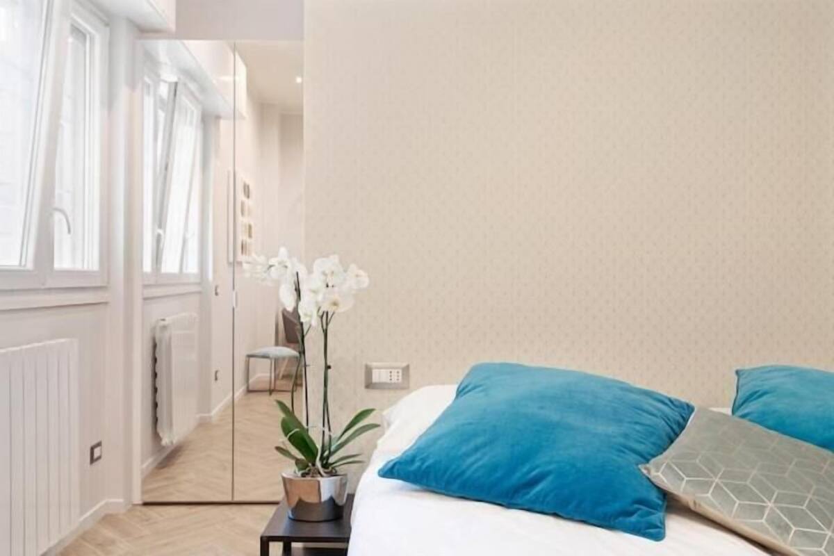 Un dormitorio con una cama, dos almohadas, una mesita de noche, una planta en maceta y una ventana.