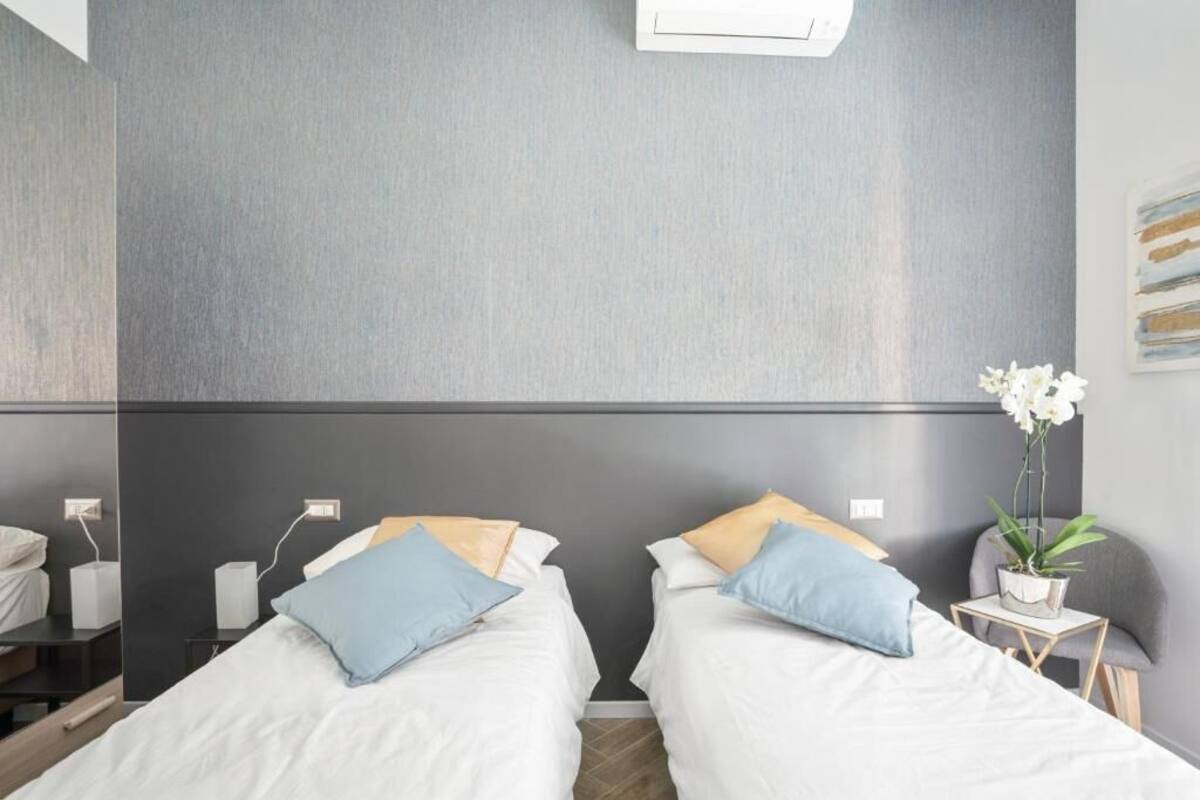 Habitación de hotel con dos camas, una mesita de noche con lámpara, una planta pequeña en maceta y un aire acondicionado instalado en la pared.