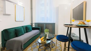 A compact living space with a green sofa, a small round table with a plant and fruit, a blue chair, and a window with sheer curtains. - Charming Studio Near Madeleine (Paris)