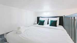 A neatly made bed with white linens and two green pillows, a white wall, and a small bedside table with a lamp. - Charming Studio Near Madeleine (Paris)