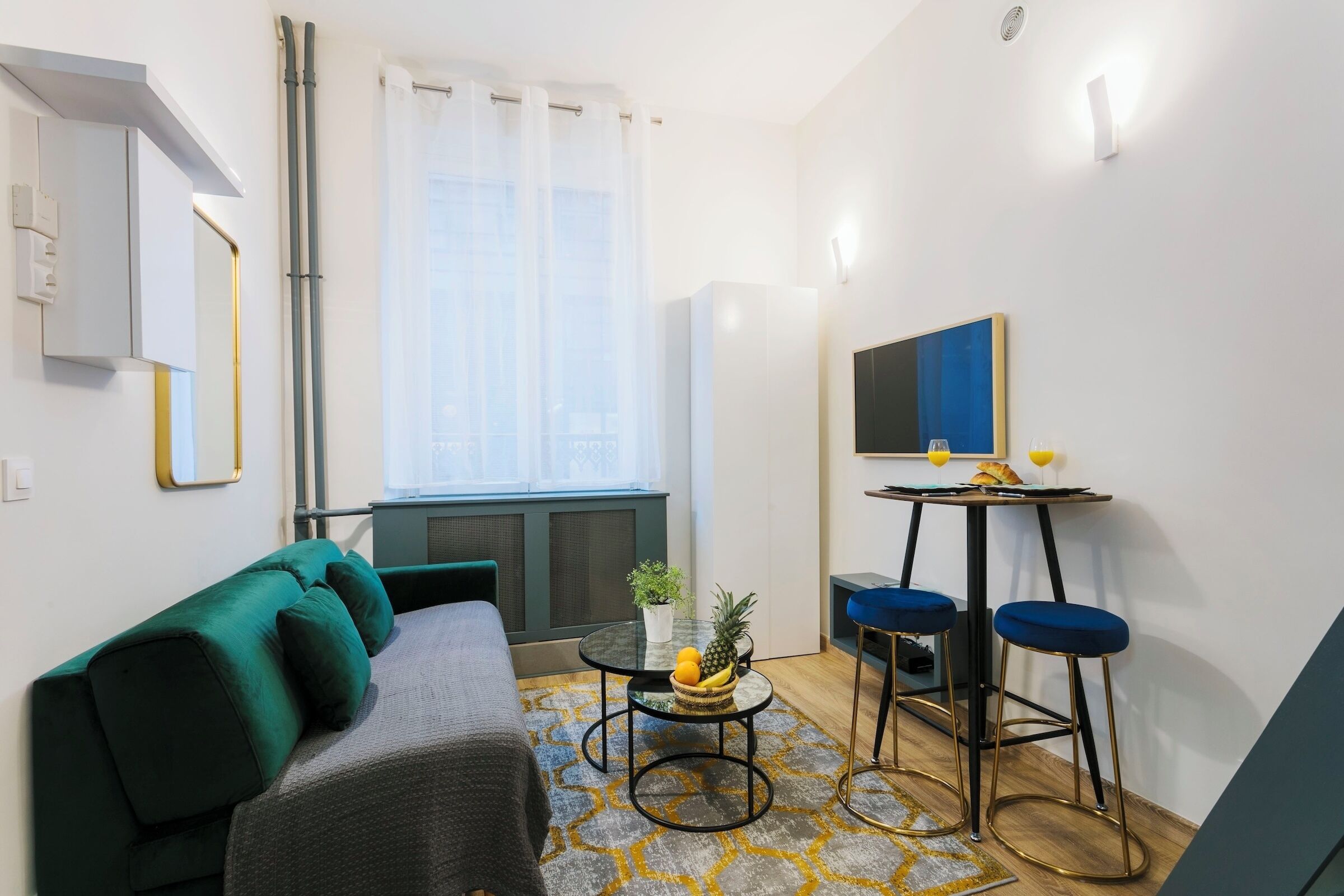 Un espacio habitable compacto con un sofá, una pequeña mesa redonda con un frutero, dos taburetes altos, un televisor y una ventana con cortinas traslúcidas.