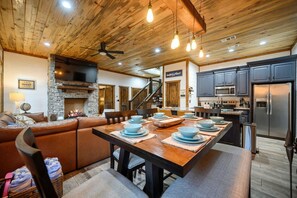 Ein gemütliches Blockhaus-Interieur mit Kamin aus Stein, einem Essbereich mit Holztisch für vier Personen und einer Küche mit modernen Geräten.