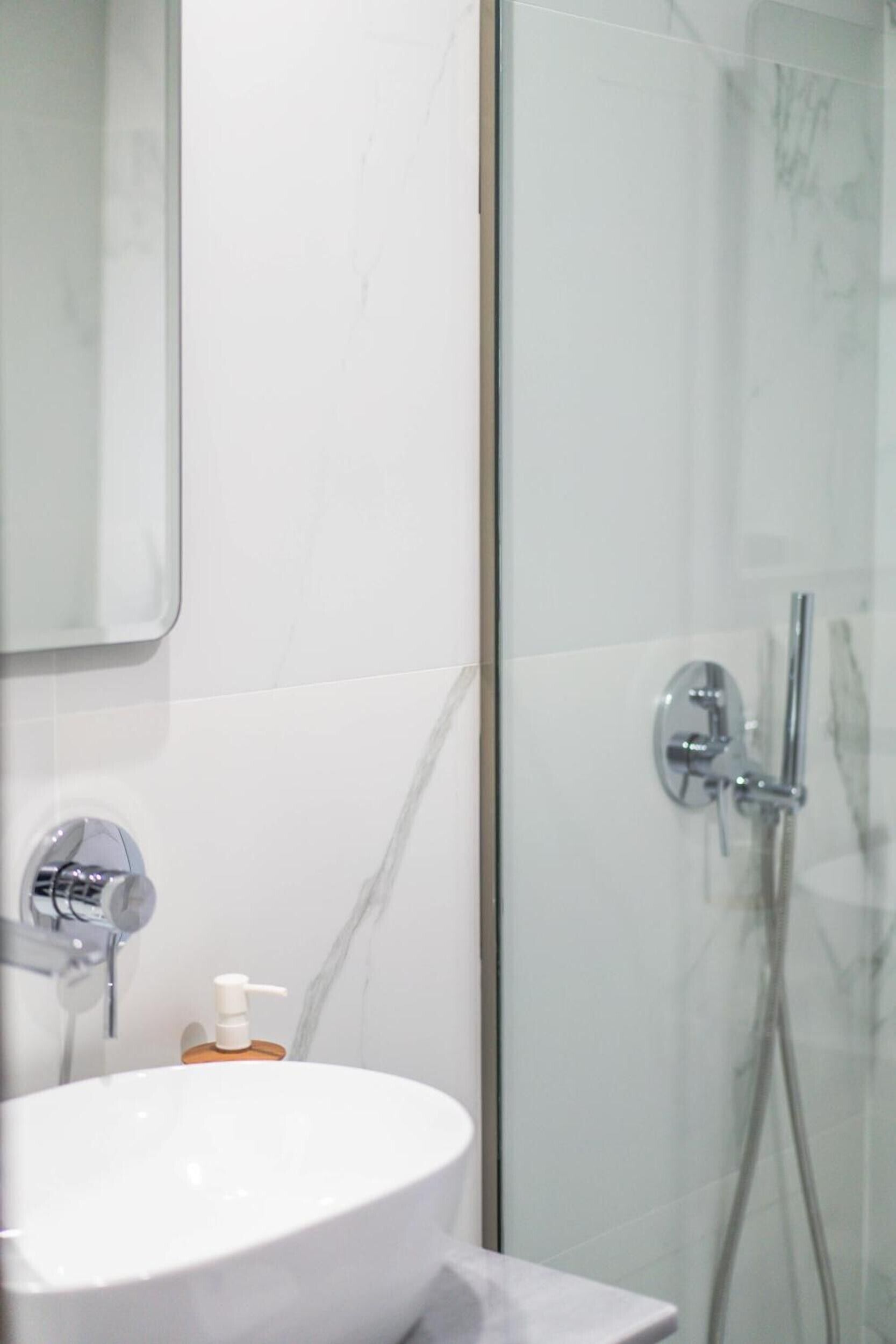 Un baño con un lavabo blanco, un espejo, cabina de ducha de vidrio y dispensador de jabón.
