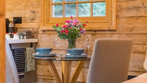 Interior de una cabaña de madera con una mesa de comedor lista para dos personas, un jarrón de flores y una ventana que da a vistas de árboles.
