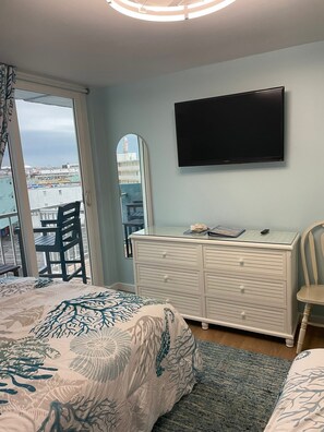 A bedroom with a bed, dresser, TV, and a view of buildings through a window. - Penthouse Beach Condo-Sunset Views & Sea Breezes! (Wildwood)