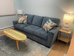 En grå soffa med mönstrade kuddar, ett träbord som soffbord, en lampa på ett sidobord och en blå matta.