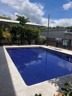 Ein rechteckiger Pool mit gefliestem Deck, umgeben von Bäumen und einem Gebäude mit Wellblechdach.