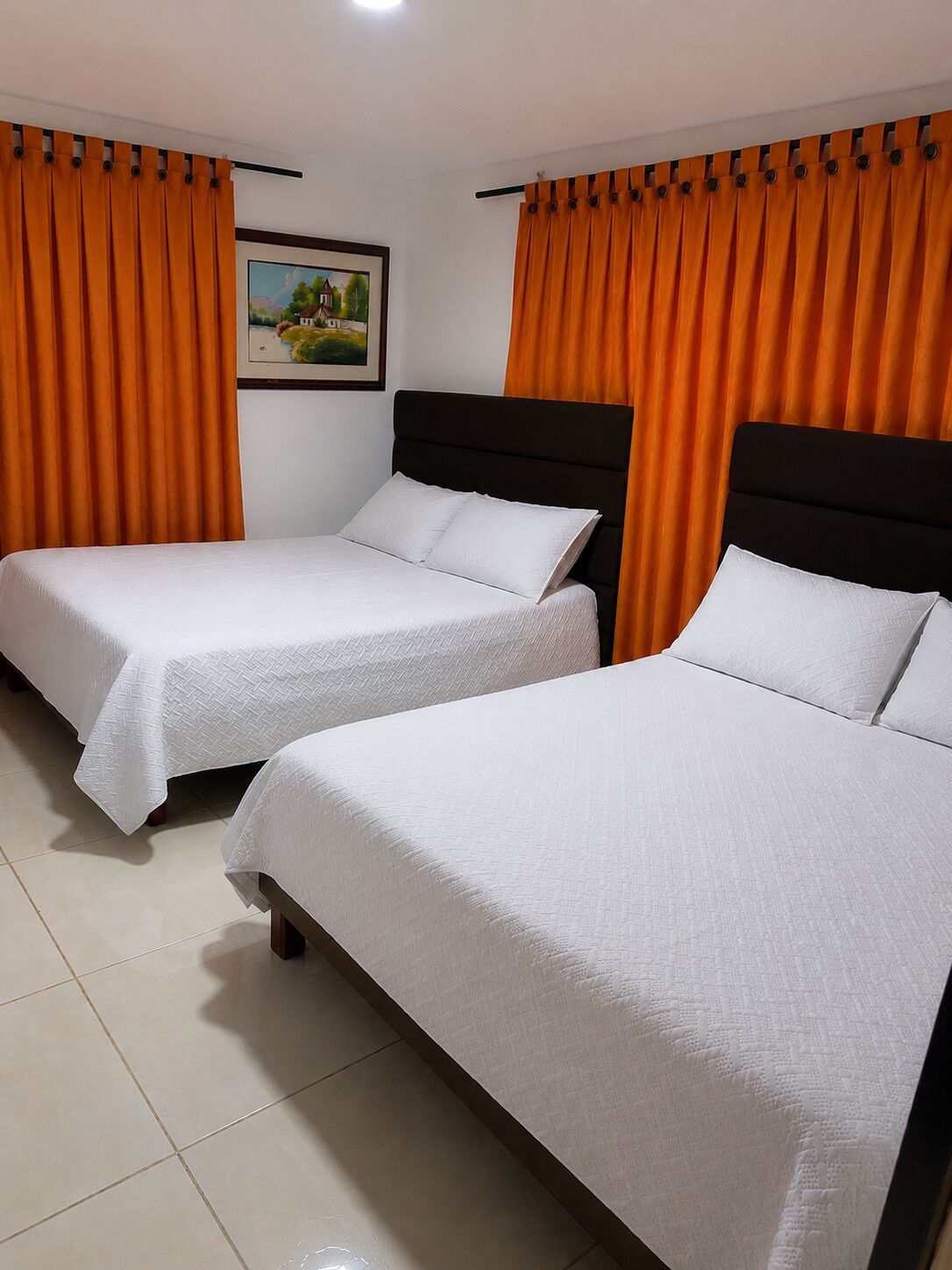 Quarto de hotel com duas camas de solteiro, cortinas laranja, um quadro emoldurado na parede e chão ladrilhado.