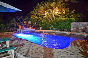A nighttime swimming pool with a water feature and a stone wall.