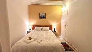 Uma cama bem arrumada com colcha, duas luminárias de cabeceira, um quadro na parede e uma porta à direita.