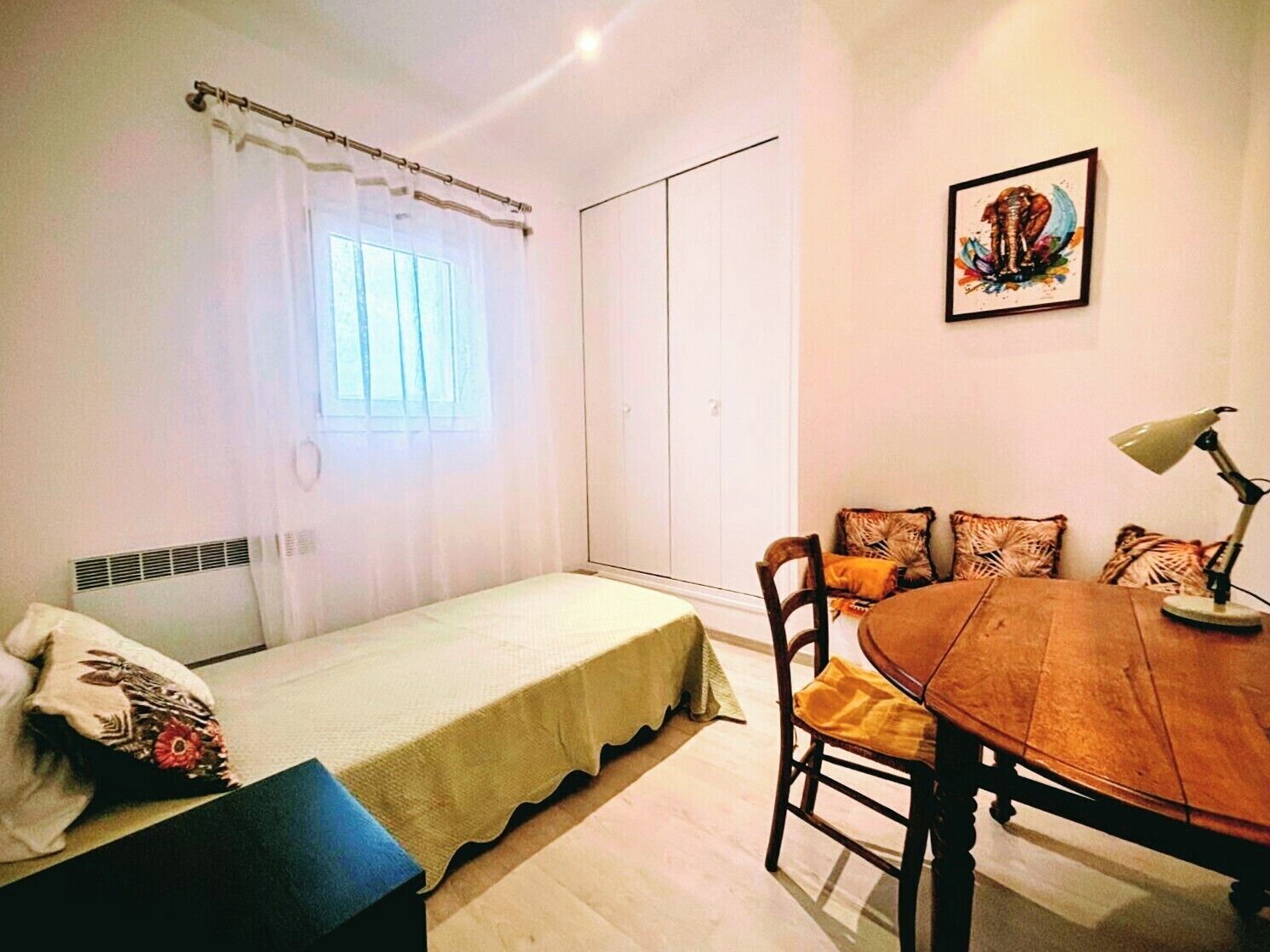 Een kleine, goed verlichte kamer met een enkel bed, een eettafel met stoelen en een raam met gordijnen.