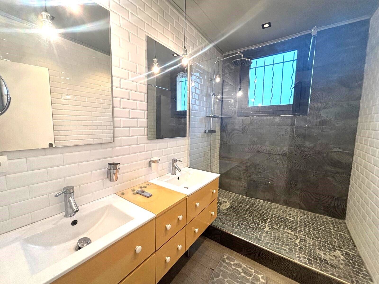 Un baño moderno con pared de azulejos blancos, tocador de madera con lavamanos y zona de ducha con mampara de vidrio.