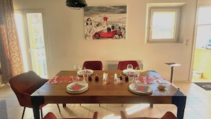 Uma sala de jantar com uma mesa de madeira posta para quatro pessoas, cadeiras vermelhas e um quadro de um carro vermelho na parede.