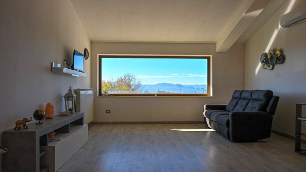 A modern living room with a large window, a black leather sofa, a small TV on a shelf, and a decorative wall piece. - Villa Donato (Filiano)