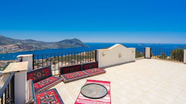 Une terrasse sur le toit offrant une vue sur la mer et les montagnes, aménagée avec des coussins aux motifs rouges et bleus et une table ronde.
