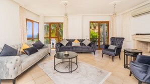 Un salon spacieux avec un canapé gris, une table basse ronde et un fauteuil gris. De grandes fenêtres sont agrémentées de rideaux, et on y trouve une cheminée ainsi qu’une vue sur la verdure à l’extérieur.