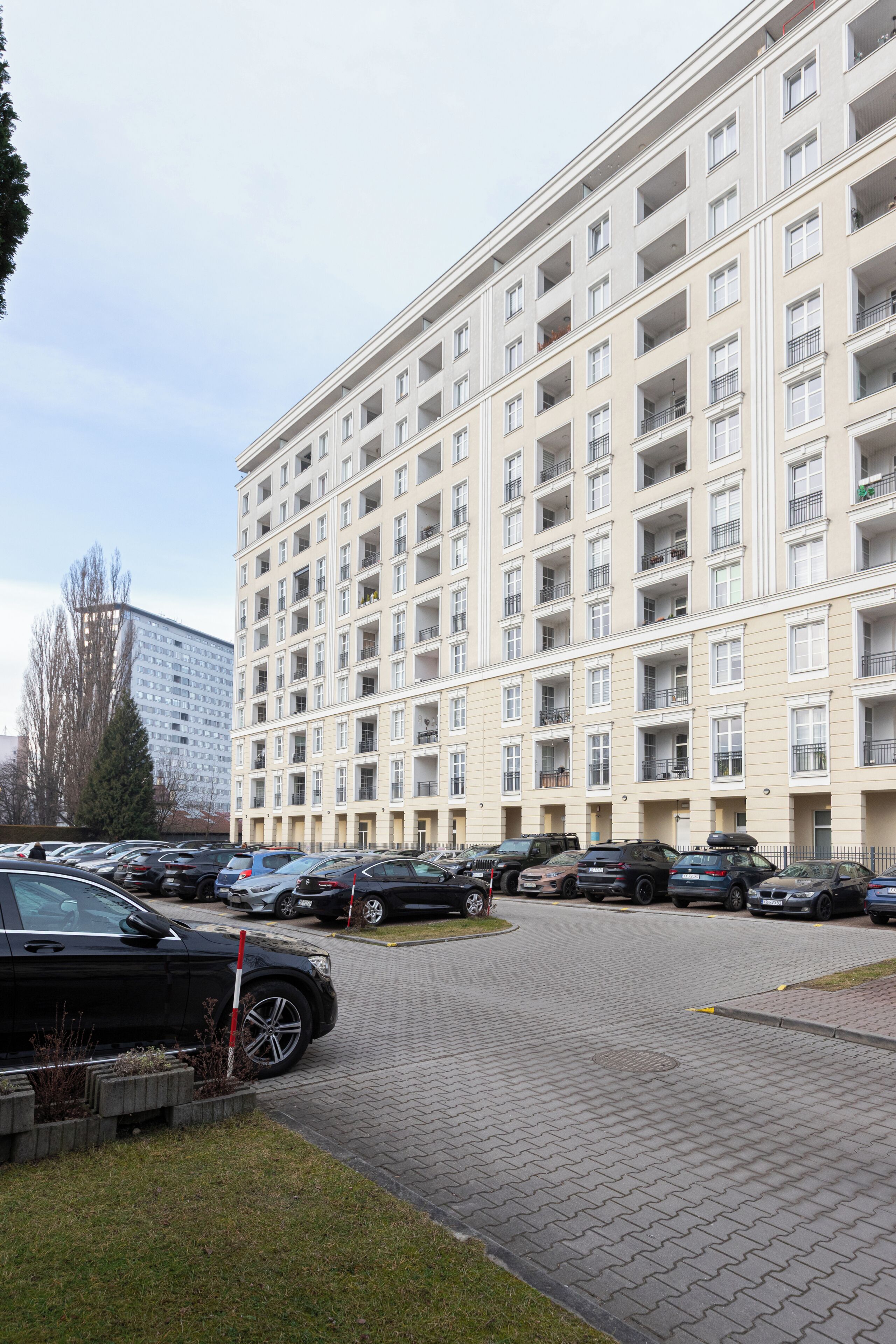 A multi-story residential building with balconies, a parking area with cars, and a clear sky.