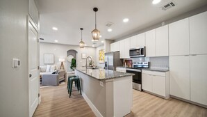 Una cocina moderna con armarios blancos, una isla central, electrodomésticos de acero inoxidable e iluminación colgante.