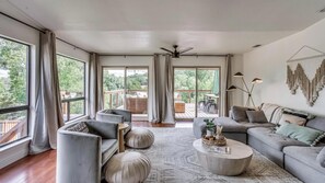 Un salon spacieux avec un grand canapé, des fauteuils et une table basse. De grandes fenêtres sont agrémentées de rideaux, un ventilateur de plafond est installé et la pièce offre une vue sur la terrasse extérieure.
