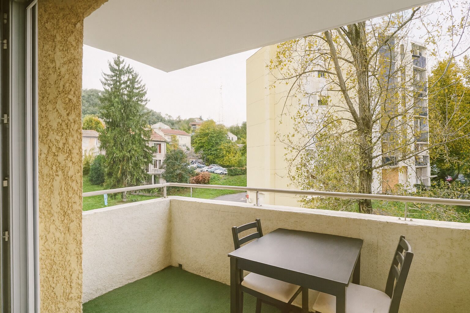 Un balcón con una mesa y sillas, vista a una zona residencial con árboles y edificios.