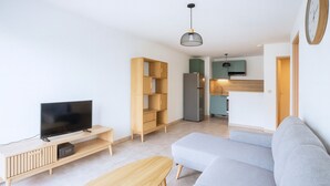 A modern living room with a flat-screen TV, wooden shelving, a grey sofa, and a kitchen area in the background.