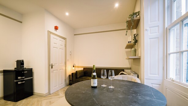 Een kamer met een ronde tafel, een fles wijn en twee glaasjes. Er is een zwart kastje met lades, een witte deur en een plank met decoratieve spullen.