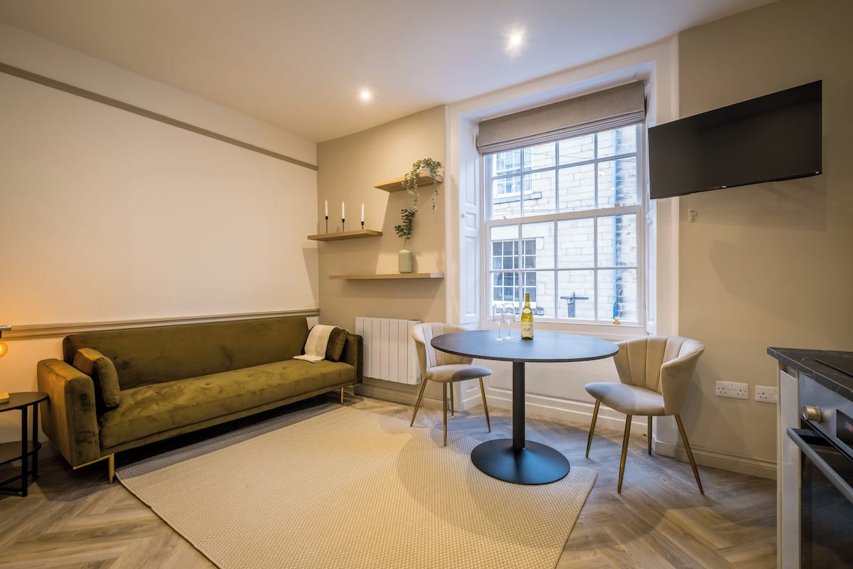 Een moderne woonkamer met een bank, een ronde tafel, stoelen en een aan de muur bevestigde televisie.