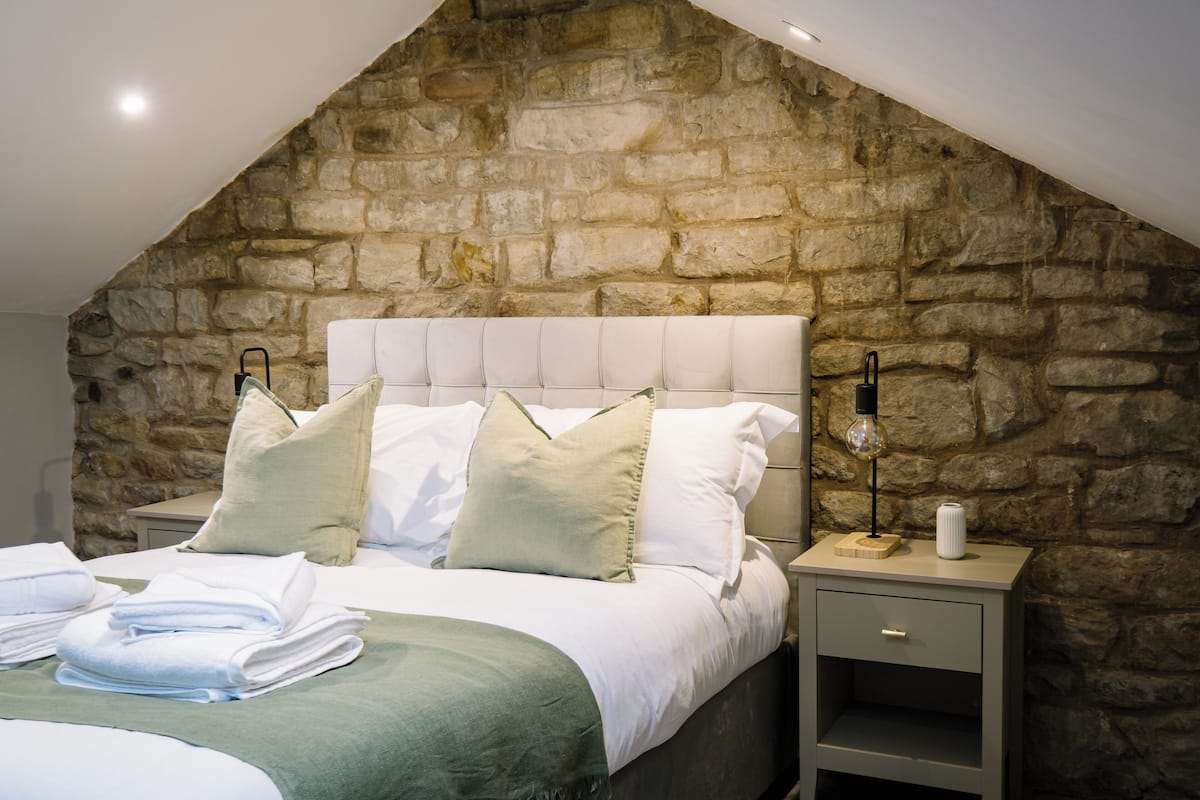 Een slaapkamer met een stenen muur, een bed met witte lakens, een nachtkastje met een lamp en een klein plankje.