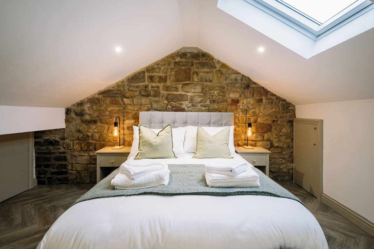 Een slaapkamer met een stenen muur, dakraam, een bed met kussens en bedlampen.