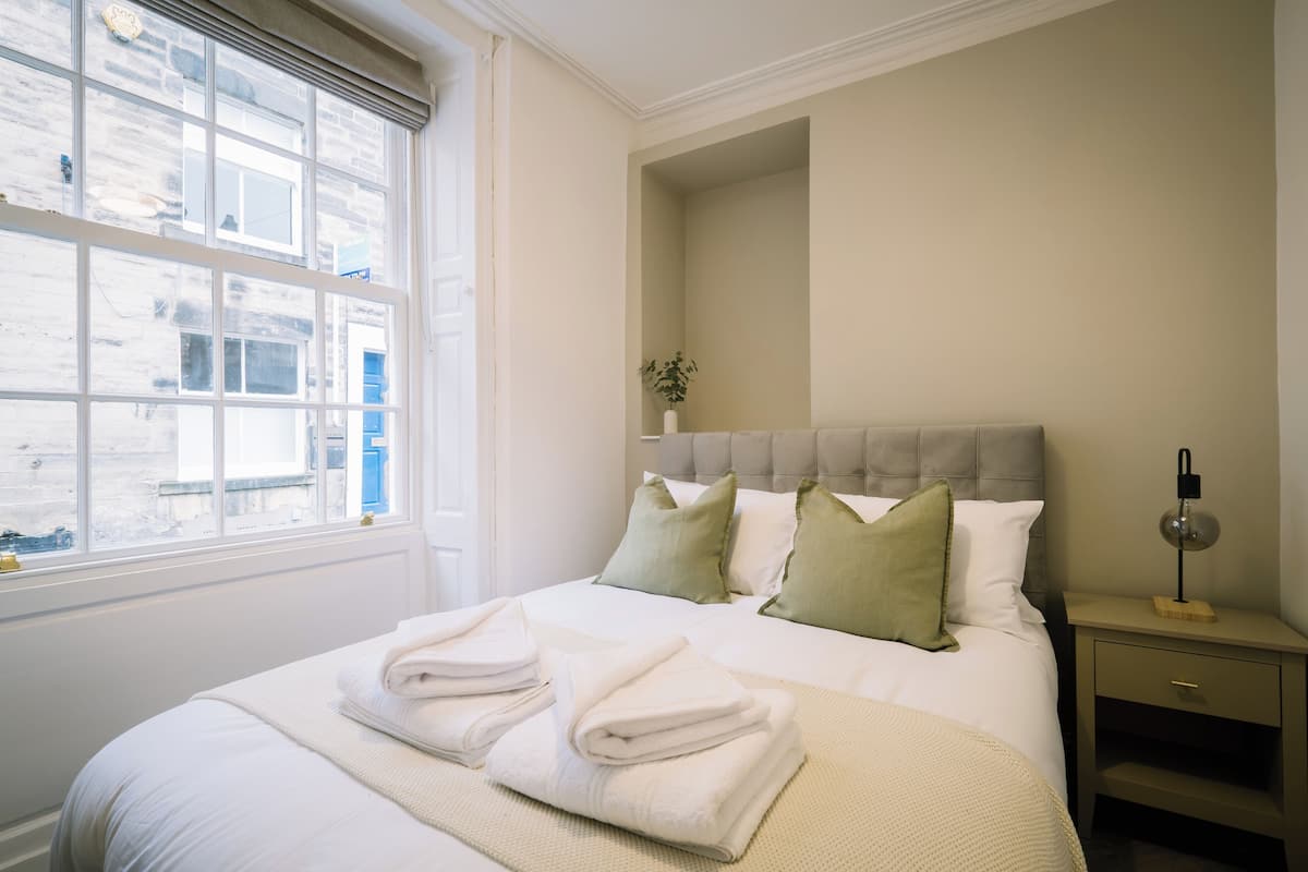 Een netjes opgemaakt bed met opgevouwen handdoeken, een nachtkastje met een lamp, en een raam met uitzicht op gebouwen.