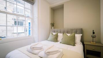A neatly made bed with folded towels, a bedside table with a lamp, and a window with a view of buildings.