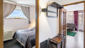 Een slaapkamer met een bed, een radiator, een raam met gordijnen, een wandlamp en uitzicht op een zithoek.