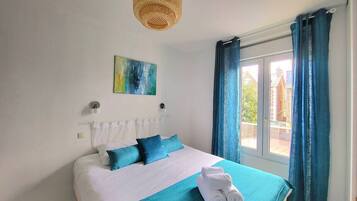 A bedroom with a bed, a window with curtains, a wall-mounted light fixture, and a painting.