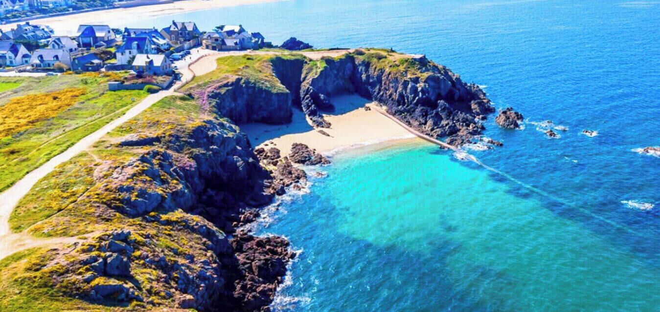 An aerial view of a coastal area with a beach, rocky cliffs, and a path leading to a cluster of houses.