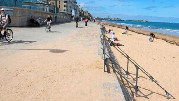Une plage avec une promenade de sable, une rambarde en métal, et des gens qui se promènent et qui sont assis.