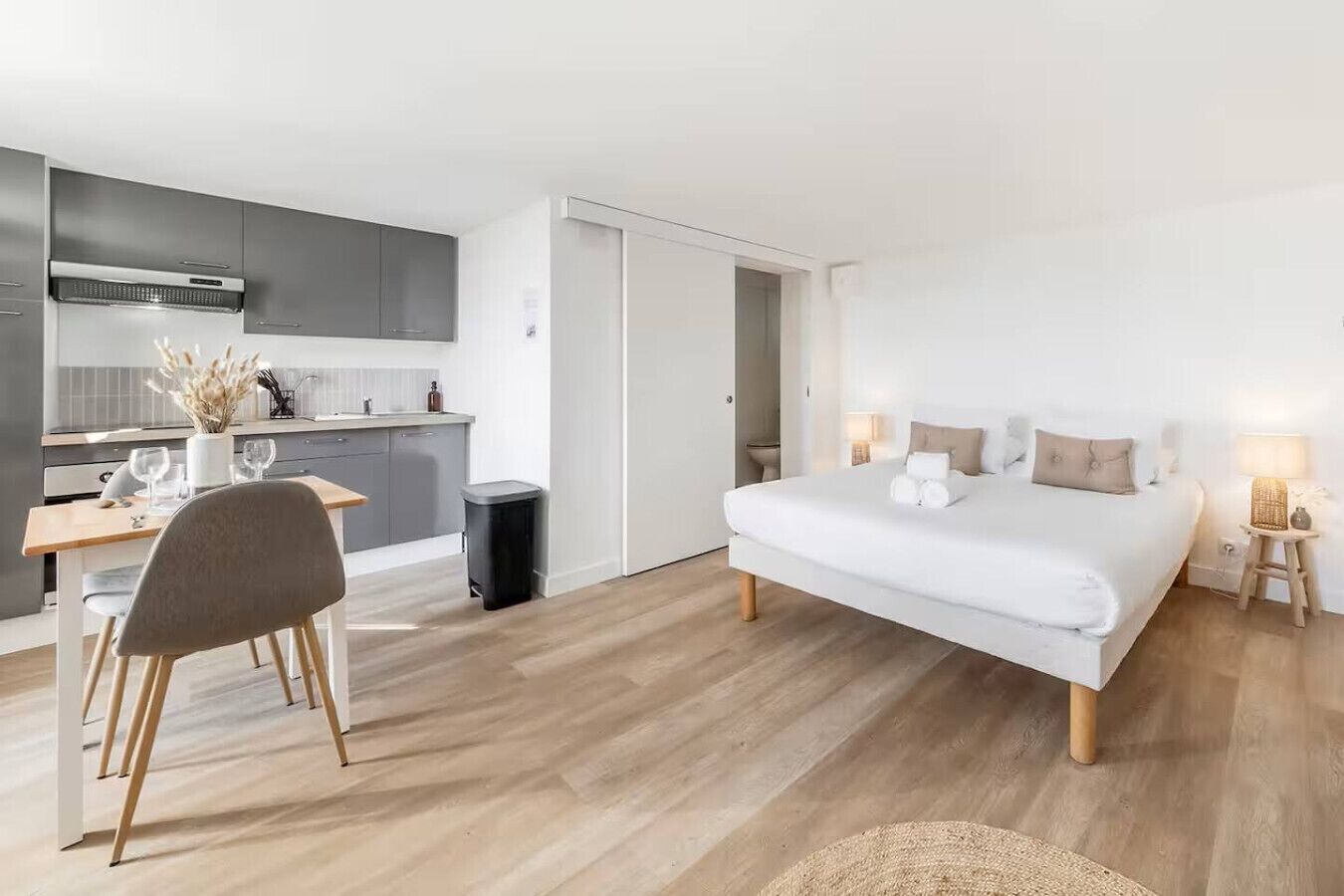 Une chambre d’hôtel moderne et compacte, équipée d’un lit, de tables de chevet, d’un petit bureau et d’une kitchenette.