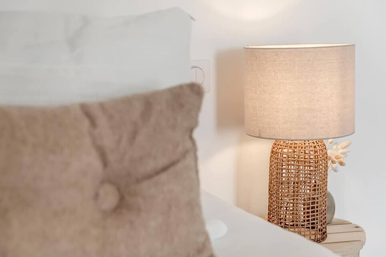 A bed with a white headboard and a brown pillow. There is a woven lamp with a beige shade on a wooden nightstand beside the bed.
