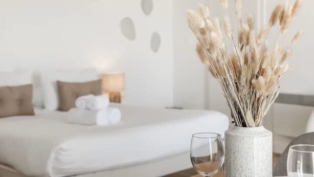 Una habitación de hotel con una cama, un florero con plumas de pampa secas y dos vasos sobre una mesa.