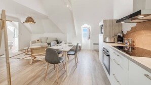 A modern kitchen with a hexagonal backsplash, a dining area with a wooden table and chairs, and a loft-style living space with a sofa and a hanging light fixture. - Parking privé - Au bord de l`eau 6 (Cancale)