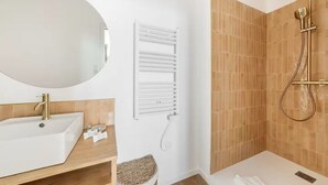 Ein modernes Badezimmer mit einem weißen Waschbecken, einem goldenen Wasserhahn und einem großen Spiegel. An der Wand ist ein Handtuchhalter angebracht und eine Dusche mit goldfarbener Duschbrause vorhanden.